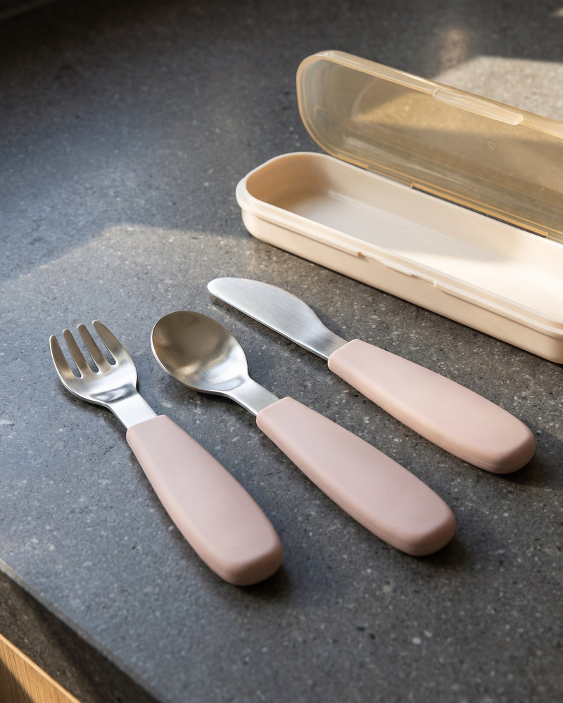 Set de cubiertos infantiles en rosa con tenedor, cuchara y cuchillo junto a su estuche sobre una encimera de piedra gris oscuro