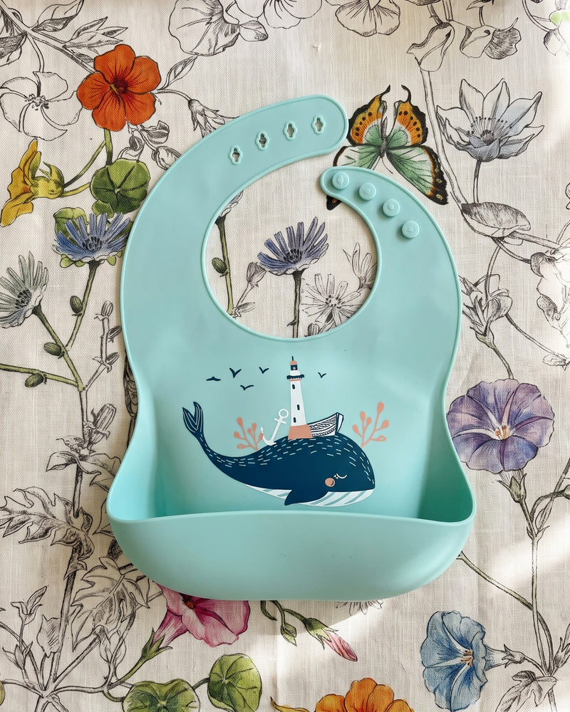 Babero de silicona verde menta con estampado de ballena y bolsillo recogemigas sobre un mantel de lino floral
